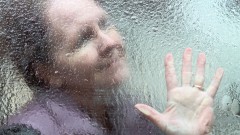 Ältere Frau blickt durch eine regennasse Fensterscheibe.