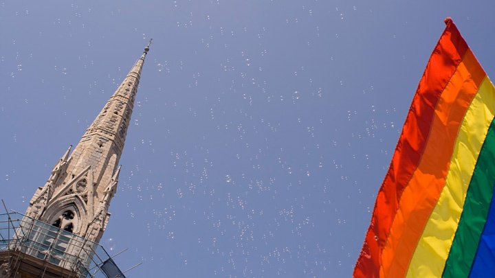 Regenbogenflagge und Kirchturm vor blauem Himmel.